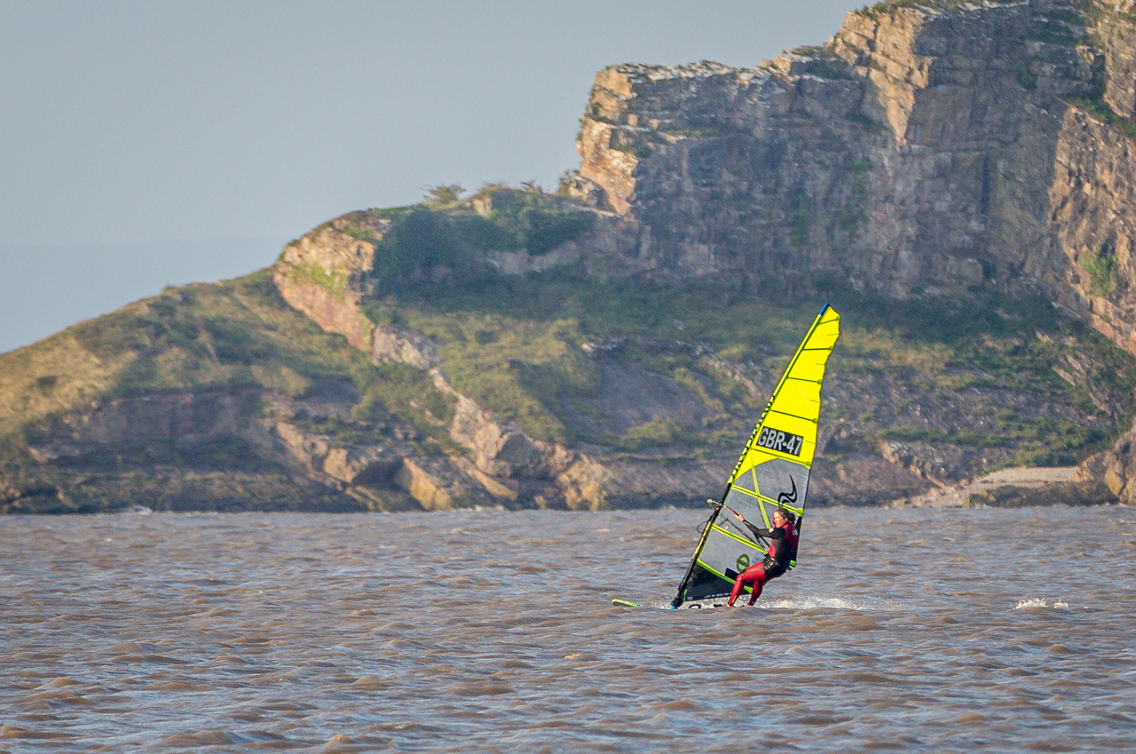 Windsurfer at sea in a black and red wetsuit and holding a yellow and black sail